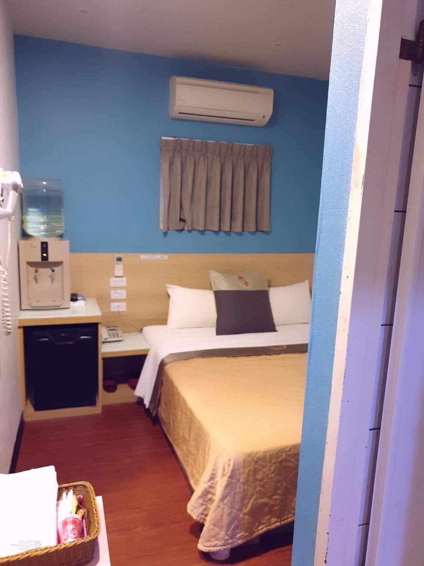 Ett hotellrum med en säng, en vattenautomat, ett litet skrivbord med en telefon och en väggmonterad luftkonditioneringsanläggning.