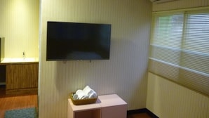 A room with a flat-screen TV mounted on the wall, a small table with a basket of towels, and a radiator with blinds.
