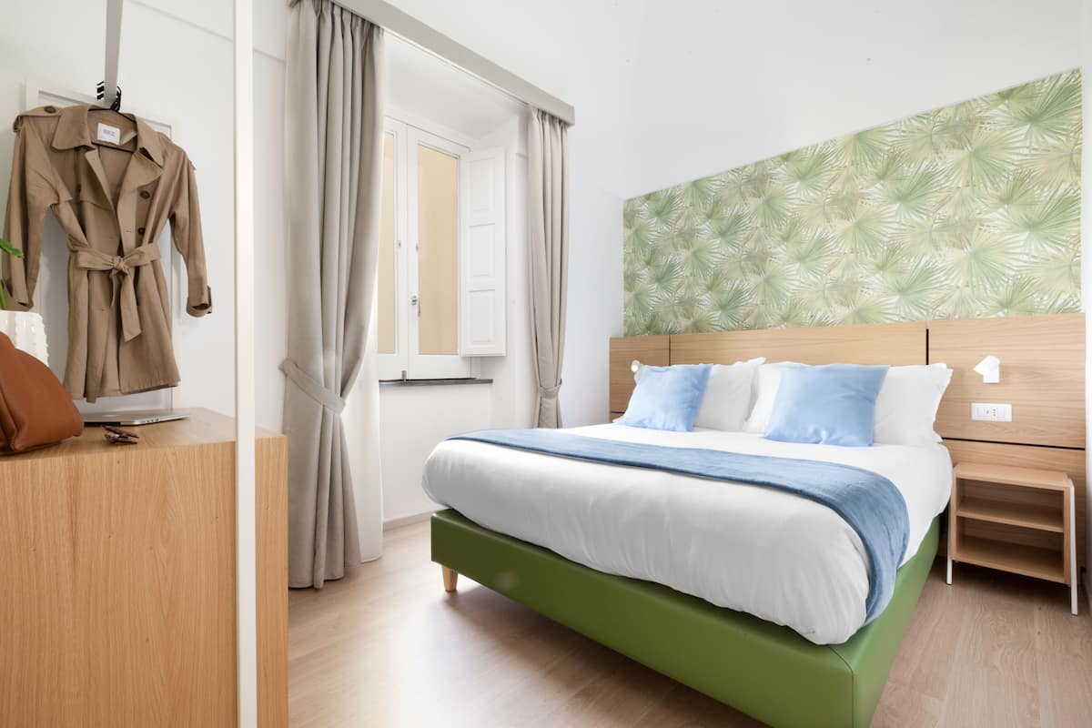 A bedroom with a bed, a wooden nightstand, a green upholstered headboard, a wooden dresser, and a coat hanging on a rack.