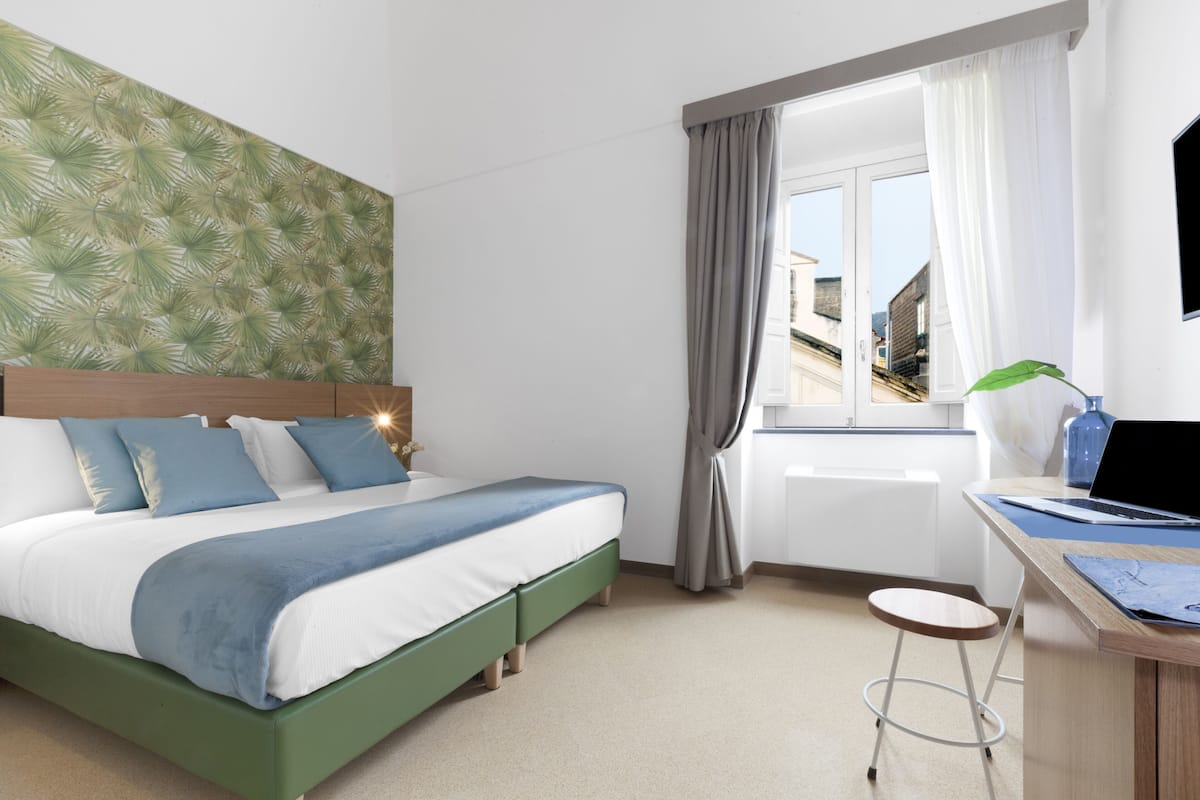 A modern hotel room with a bed, a desk with a laptop, a window with curtains, and a view of buildings outside.