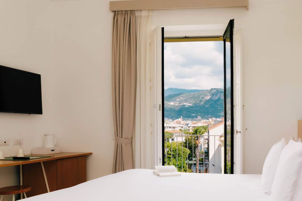 A hotel room with a bed, a desk, a chair, a TV, and a window with a view of a city and mountains.