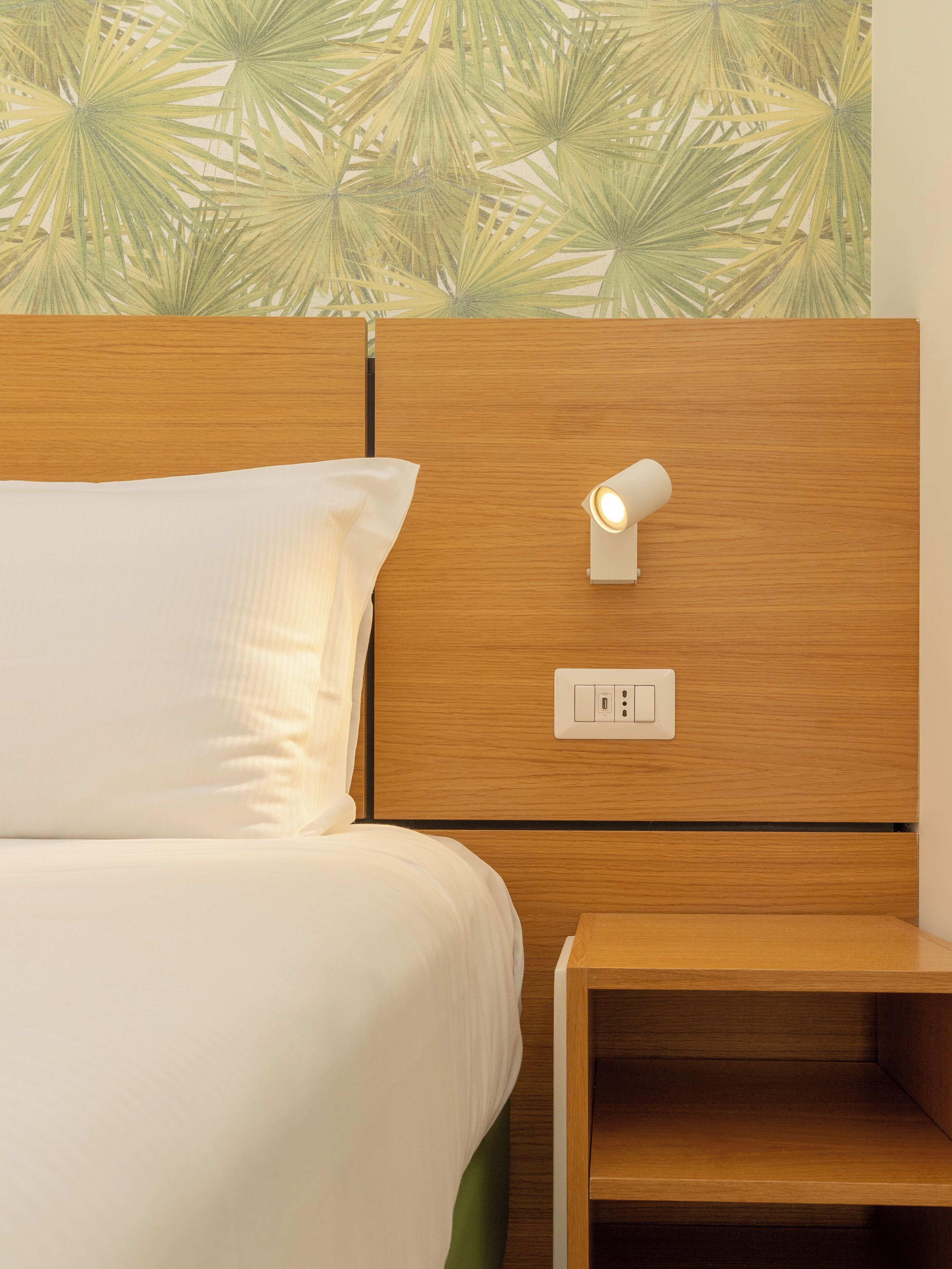Een netjes opgemaakt bed met een wit kussen en een houten hoofdbord, met een wandlamp.