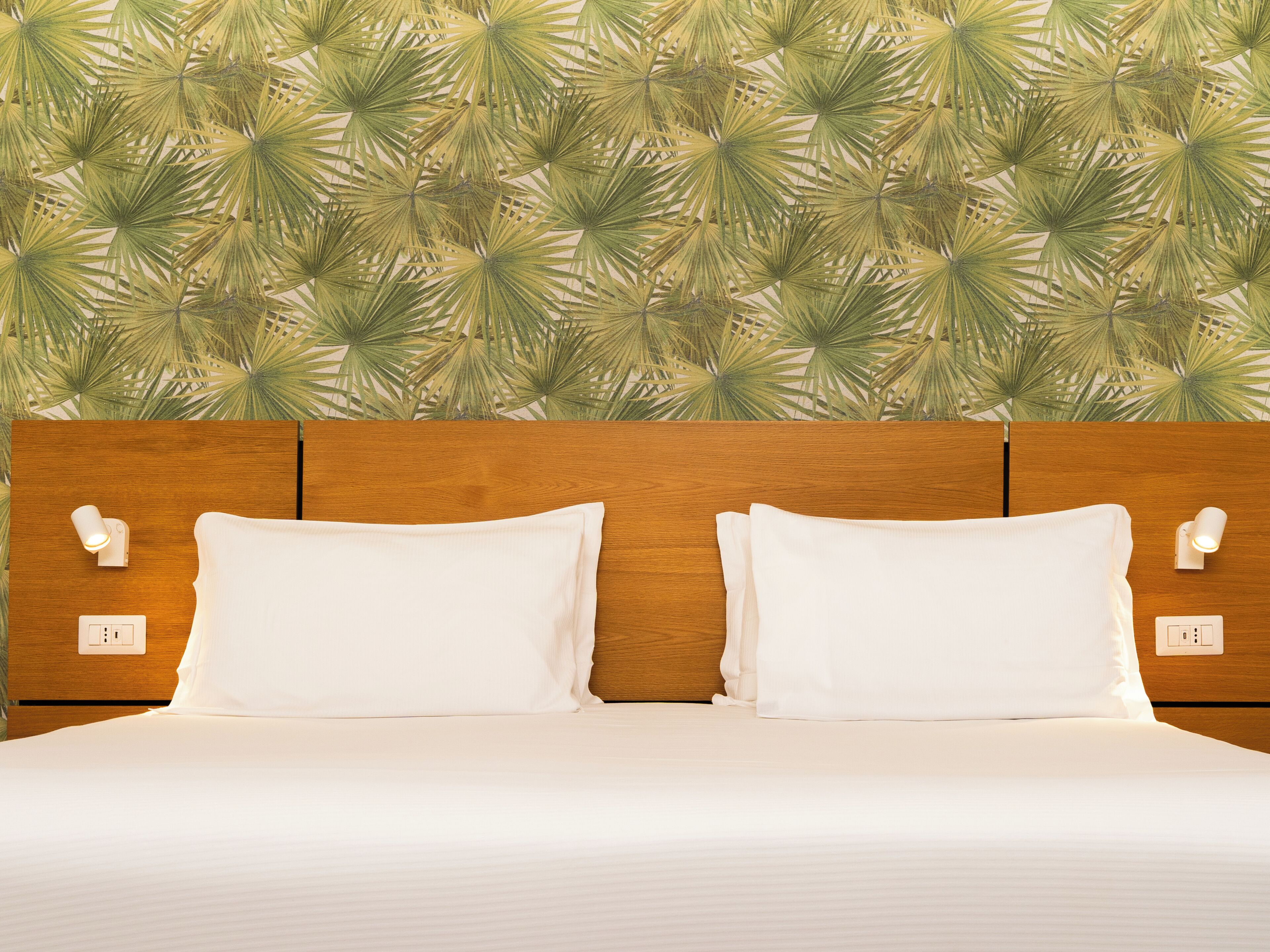 Habitación de hotel con cabecera de madera, dos almohadas blancas y pared con estampado de hojas tropicales.
