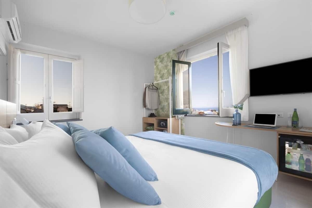 A hotel room with a large bed, a desk with a laptop, a TV mounted on the wall, and a window with curtains.