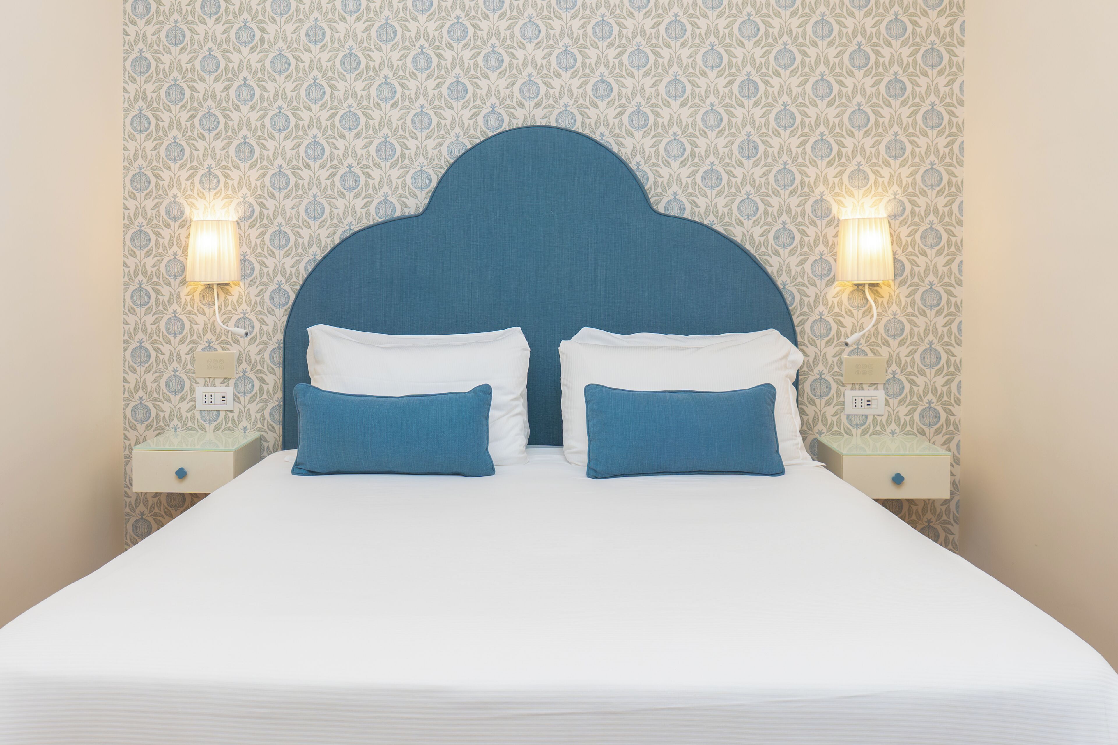 Una cama con cabecera azul, dos mesitas de noche con lámparas y un papel tapiz estampado.