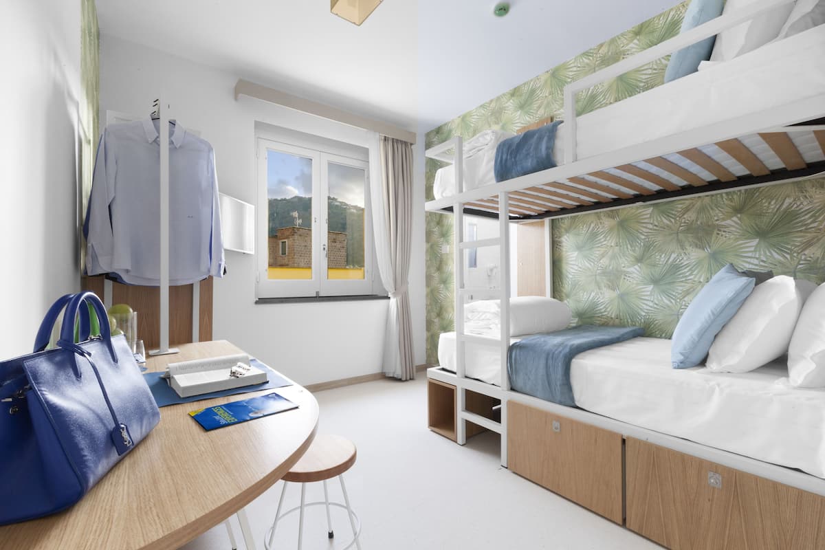A room with bunk beds, a desk with a chair, a blue bag, and a window with curtains.