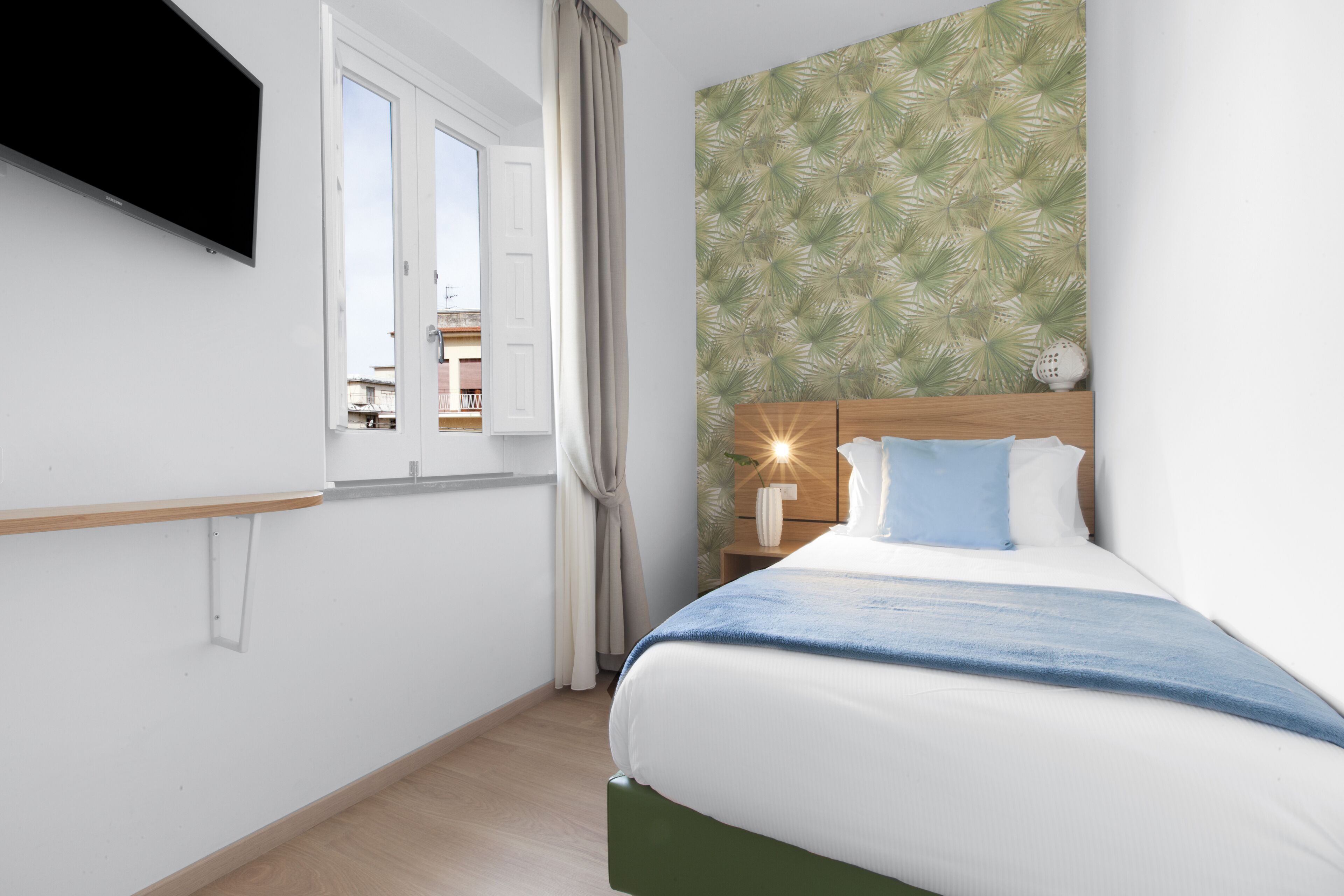 Un dormitorio con cama, mesita de noche, televisor en la pared, ventana con cortinas y un papel tapiz estampado.