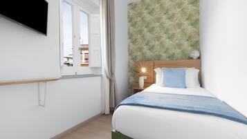 Un dormitorio con cama, mesita de noche, televisor en la pared, ventana con cortinas y un papel tapiz estampado.