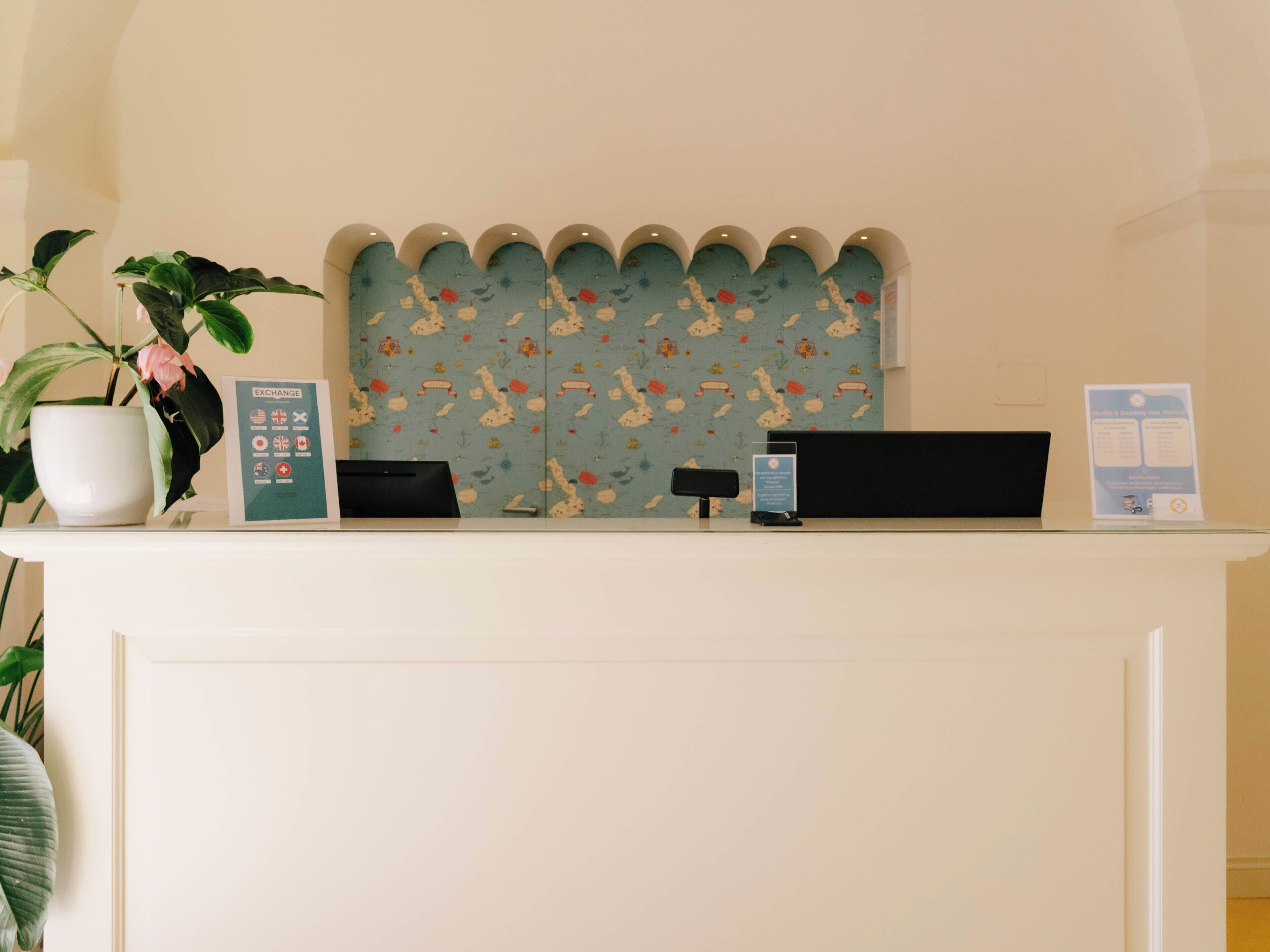 Un mostrador de recepción con un fondo estampado de flores, una planta en maceta y carteles informativos.