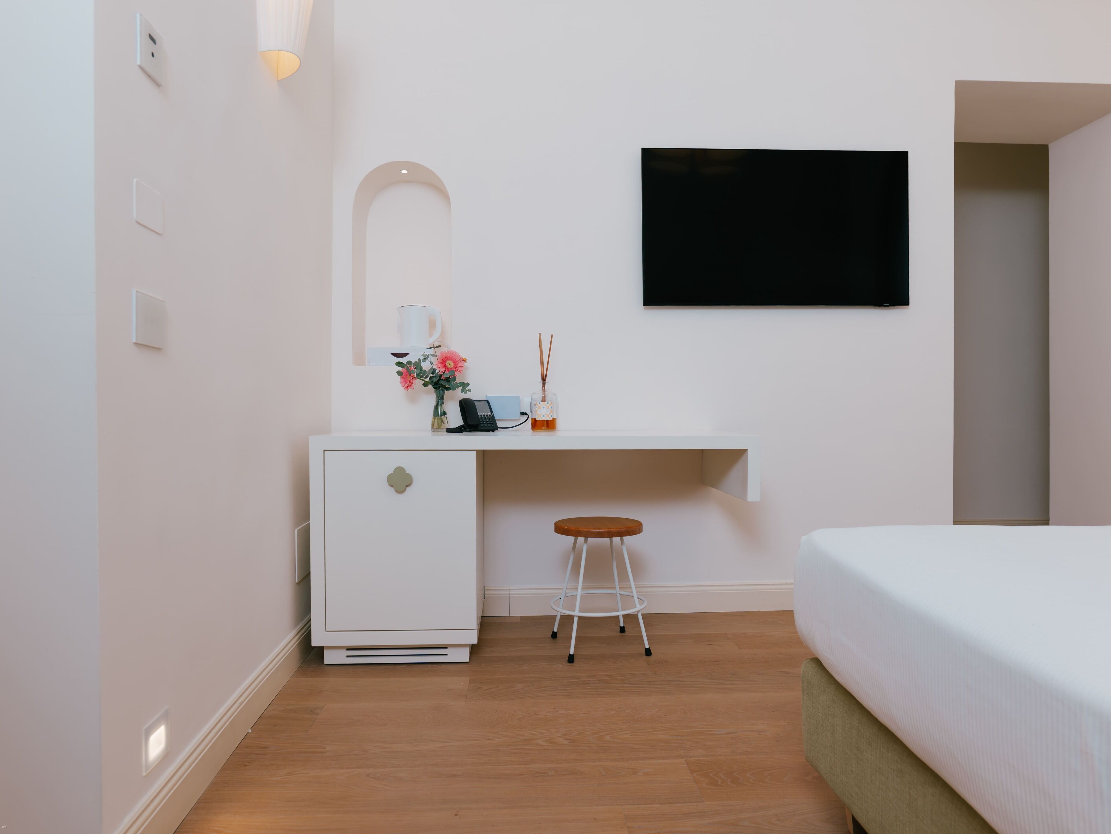 A compact hotel room with a bed, a small desk with a chair, a TV mounted on the wall, and a compact refrigerator.