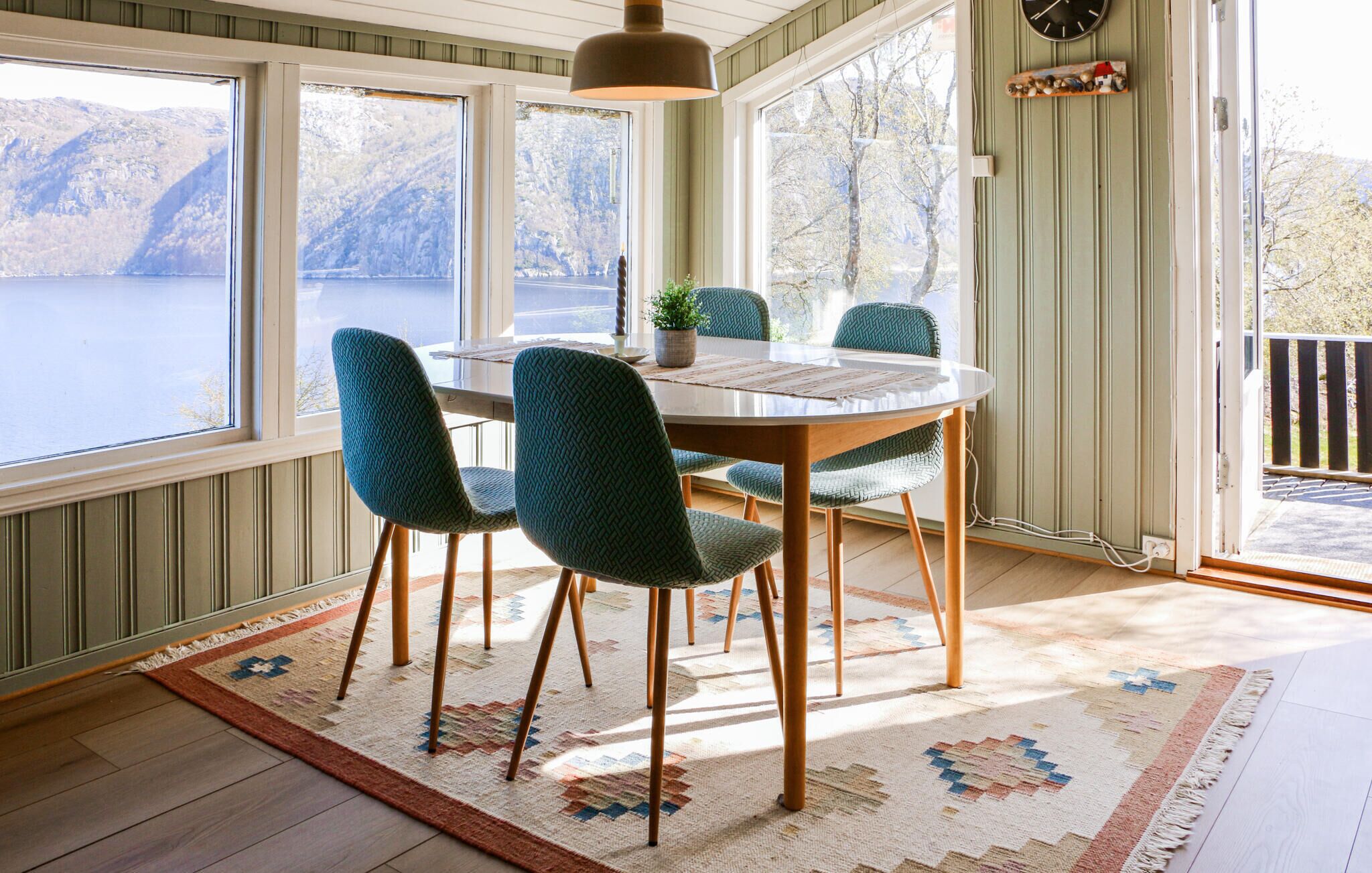 Un comedor con una mesa redonda, cuatro sillas, una lámpara colgante y vistas a un lago a través de amplios ventanales.