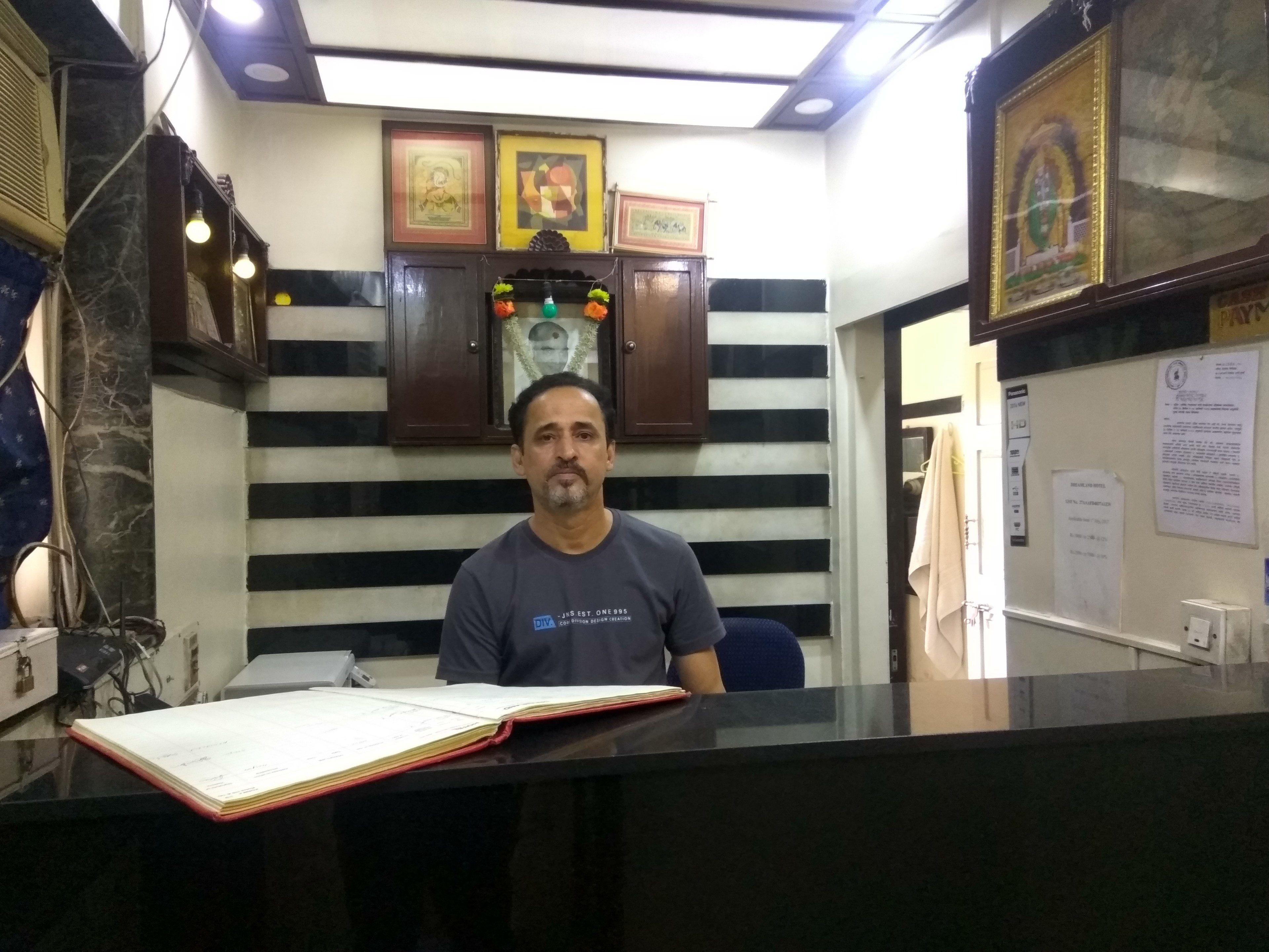 Un hombre sentado en un escritorio con un libro abierto frente a él. Hay cuadros en la pared detrás y un tablón de anuncios con papeles.