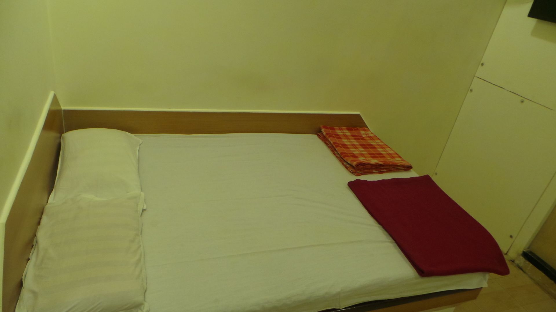 A small, single-bed room with a plain white mattress, a red blanket, and a checkered blanket on the bed.