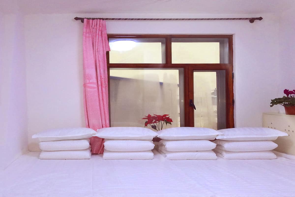 A room with a bed covered in white linens, a window with a wooden frame, and a pink curtain.