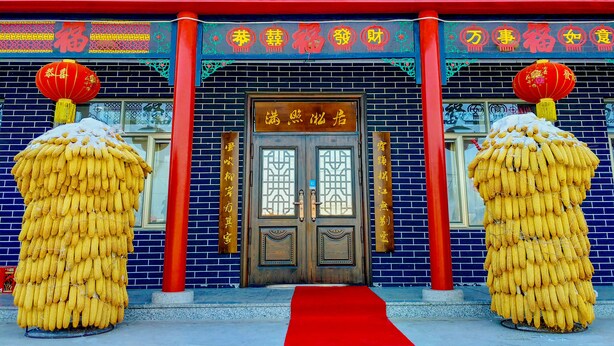 A traditional building with a red and blue facade, a wooden door with a sign above it, and two large corn sculptures on either side of the entrance.