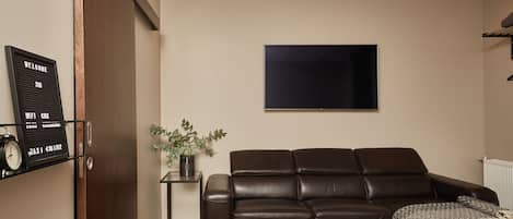 Una sala de estar moderna con un sofá de cuero, un televisor de pantalla plana, una mesita auxiliar con una planta y un letrero de bienvenida colgado en la pared.