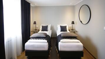 A hotel room with two single beds, each with a pillow and a blanket, a bedside table with a lamp, and a large mirror on the wall.