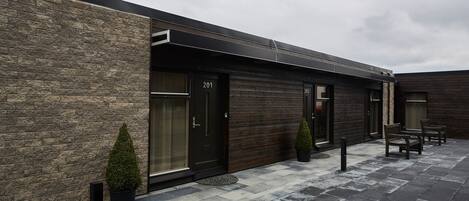 A modern building with numbered doors, a paved outdoor area, and outdoor seating.