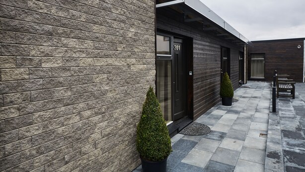 A modern building with a stone facade, a black door marked '201', and a small garden area with a bench and a potted tree. - Keflavik Micro Suites (Reykjanesbær)
