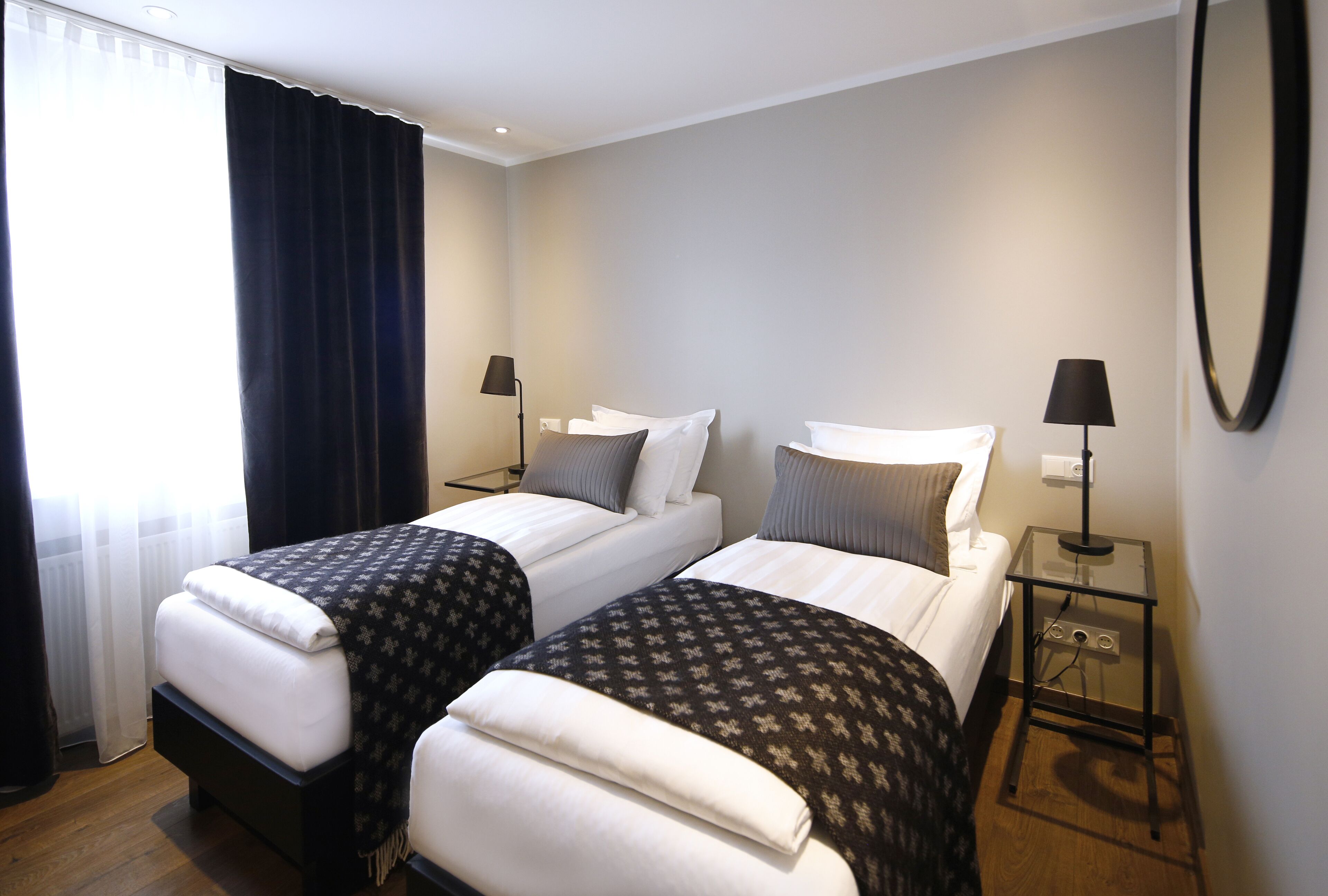 A hotel room with two single beds, a bedside table with a lamp, a mirror, and a window with curtains.