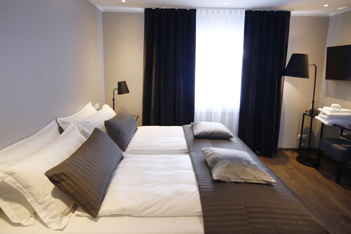 A double bed with white and brown bedding, a bedside table with a lamp, and a view of the outdoors through large windows with dark curtains.