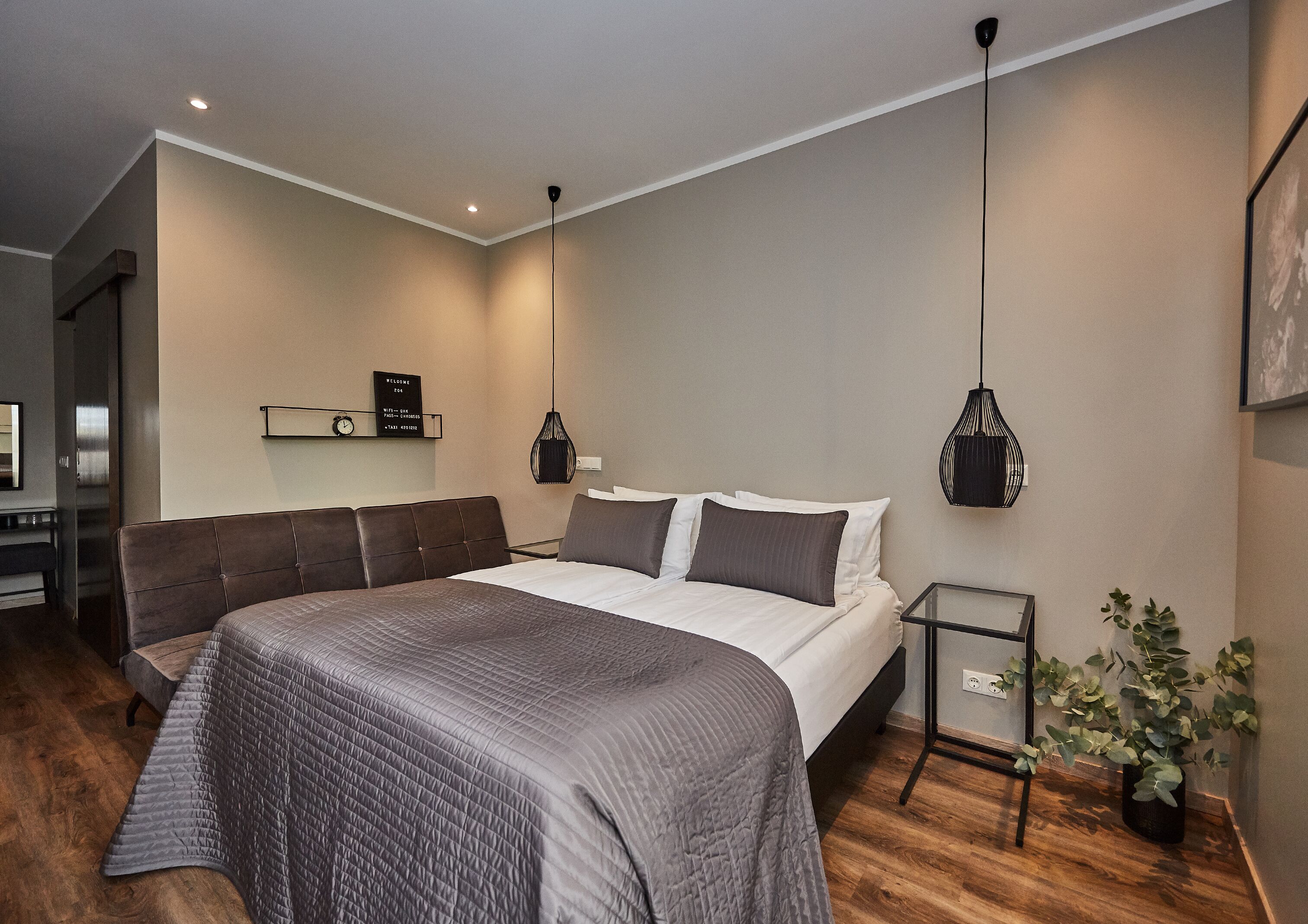 A modern hotel room with a large bed, a bedside table, a small desk, and a potted plant.