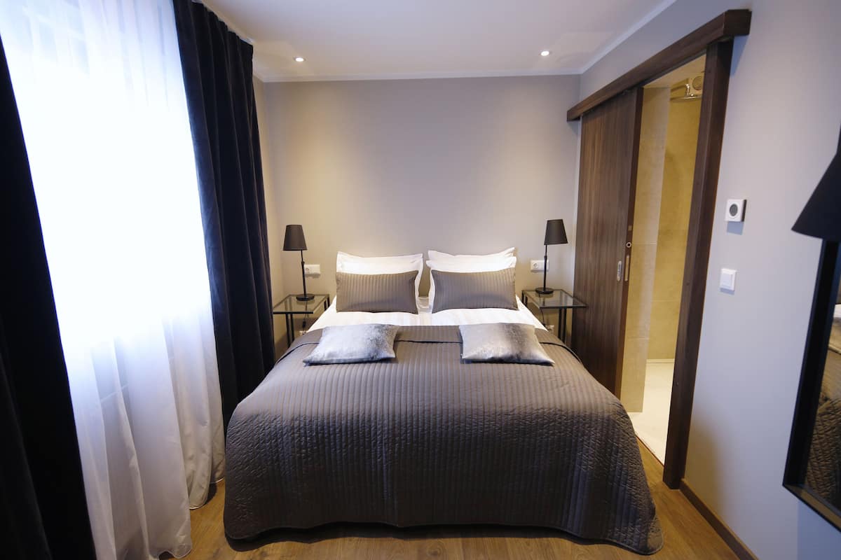 A neatly made bed with a dark bedspread and two pillows, flanked by two bedside tables with lamps, in a room with a wooden floor and a sliding door.
