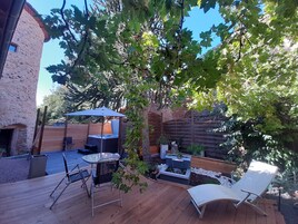 Una terraza de madera con mobiliario exterior, un jacuzzi y una pérgola con un paraguas azul.