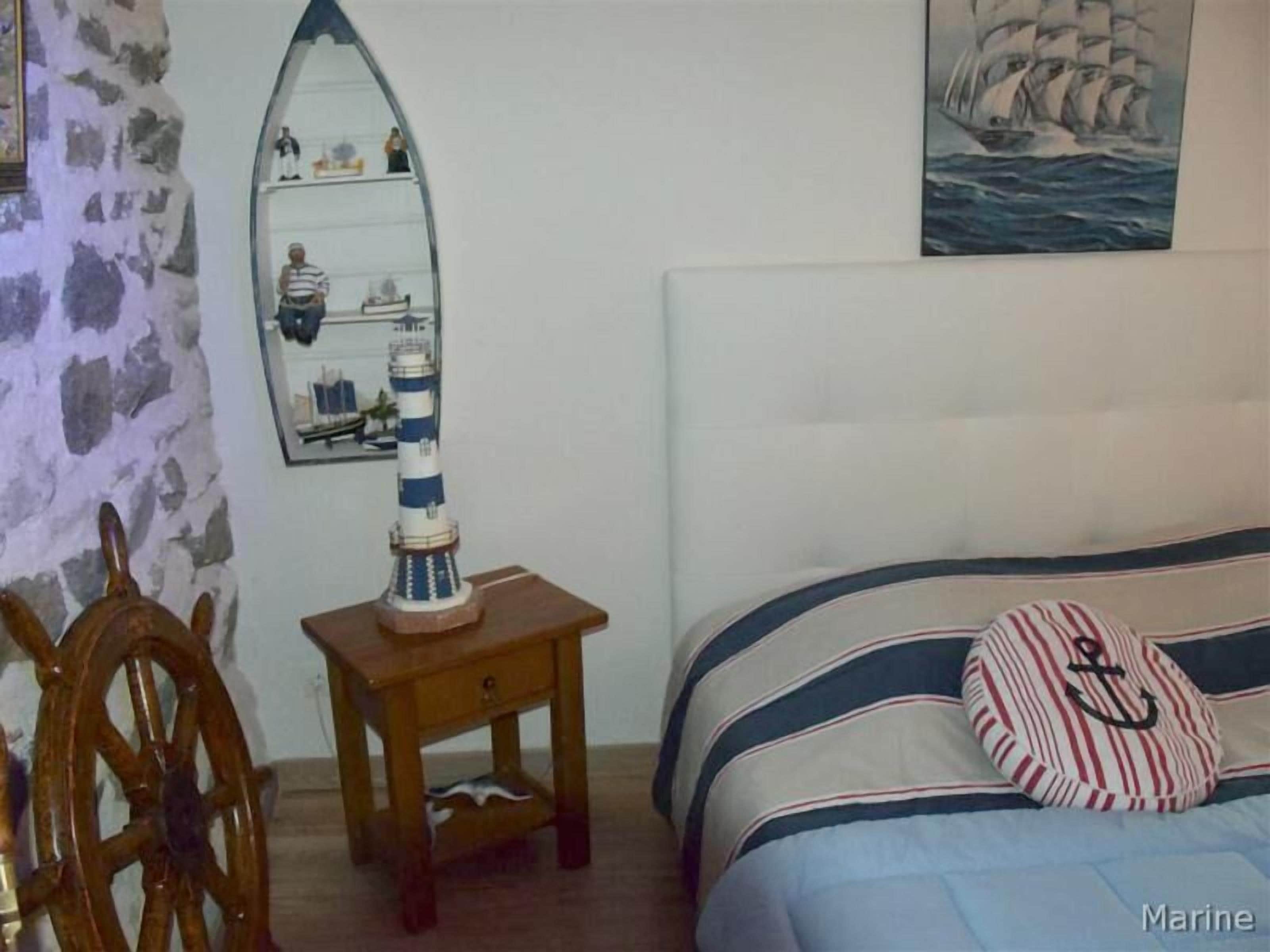 A bedroom with a bed, a wooden nightstand with a model ship, a wooden ship's wheel, and a painting of a sailing ship.
