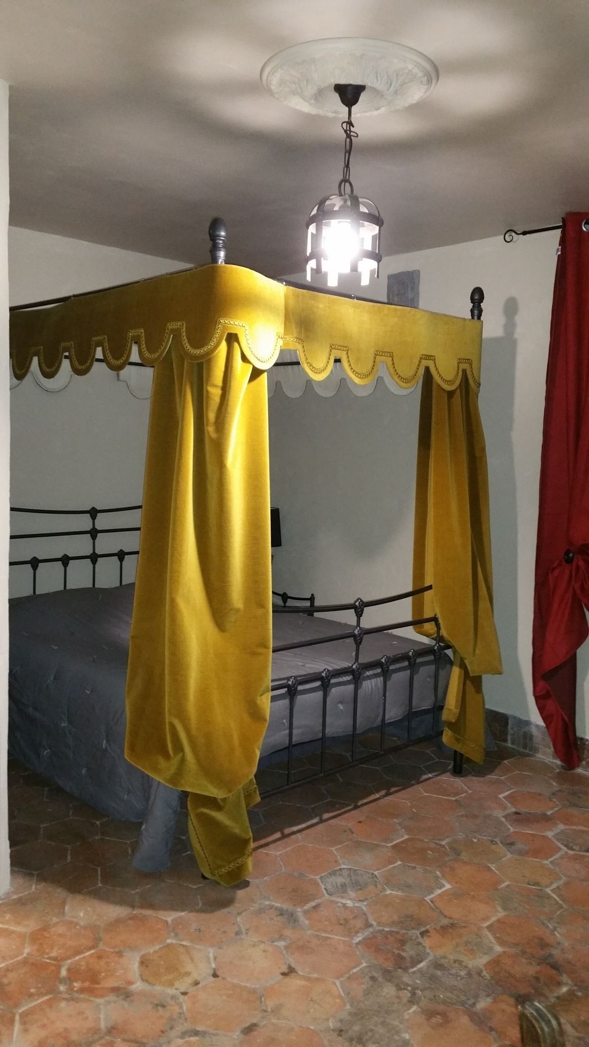 Une chambre avec un lit placé sous un dais jaune, un sol carrelé en pierre et un luminaire au plafond.