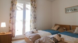 Ein Schlafzimmer mit zwei Betten, einem Fenster mit blumigen Vorhängen, einer Nachttischlampe und einem gerahmten Bild an der Wand.