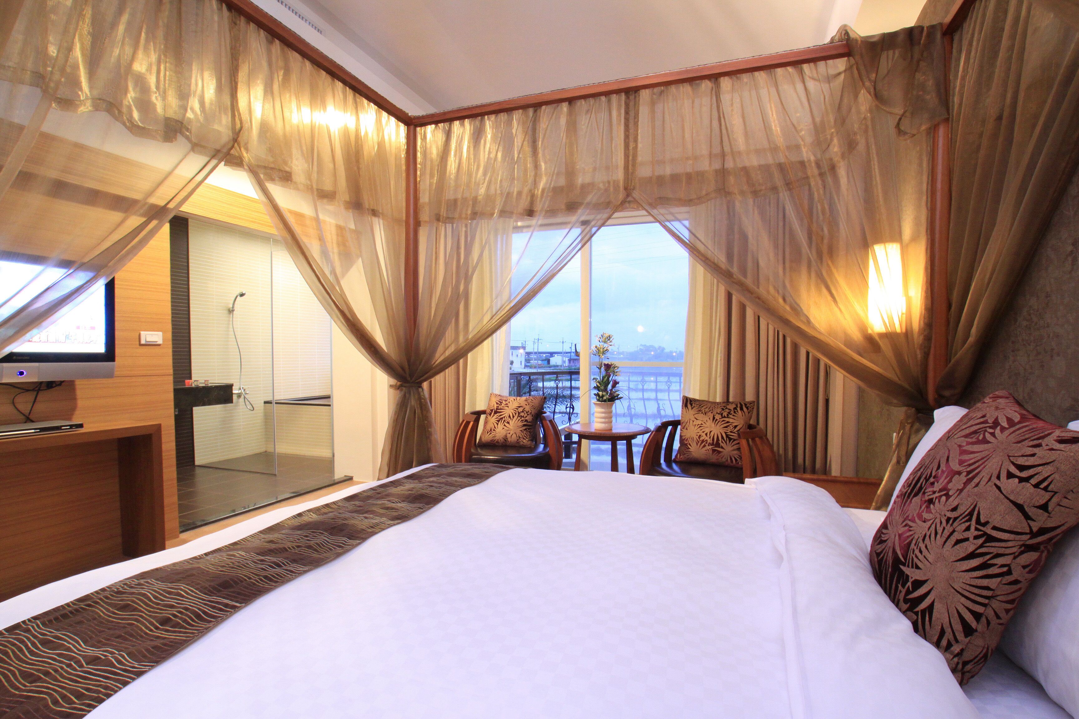 A hotel room with a large bed, a TV, a glass-enclosed shower area, and a view of the cityscape.