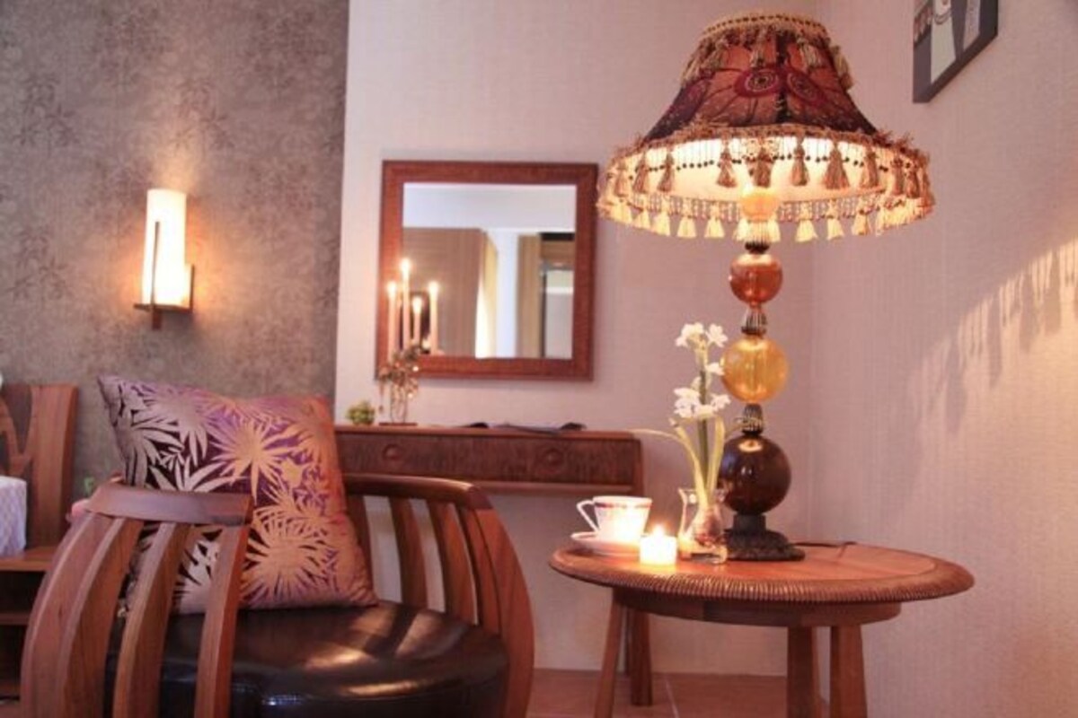 A cozy seating area with a wooden chair and a cushioned bench, a table with a lamp and candles, and a wall-mounted mirror.