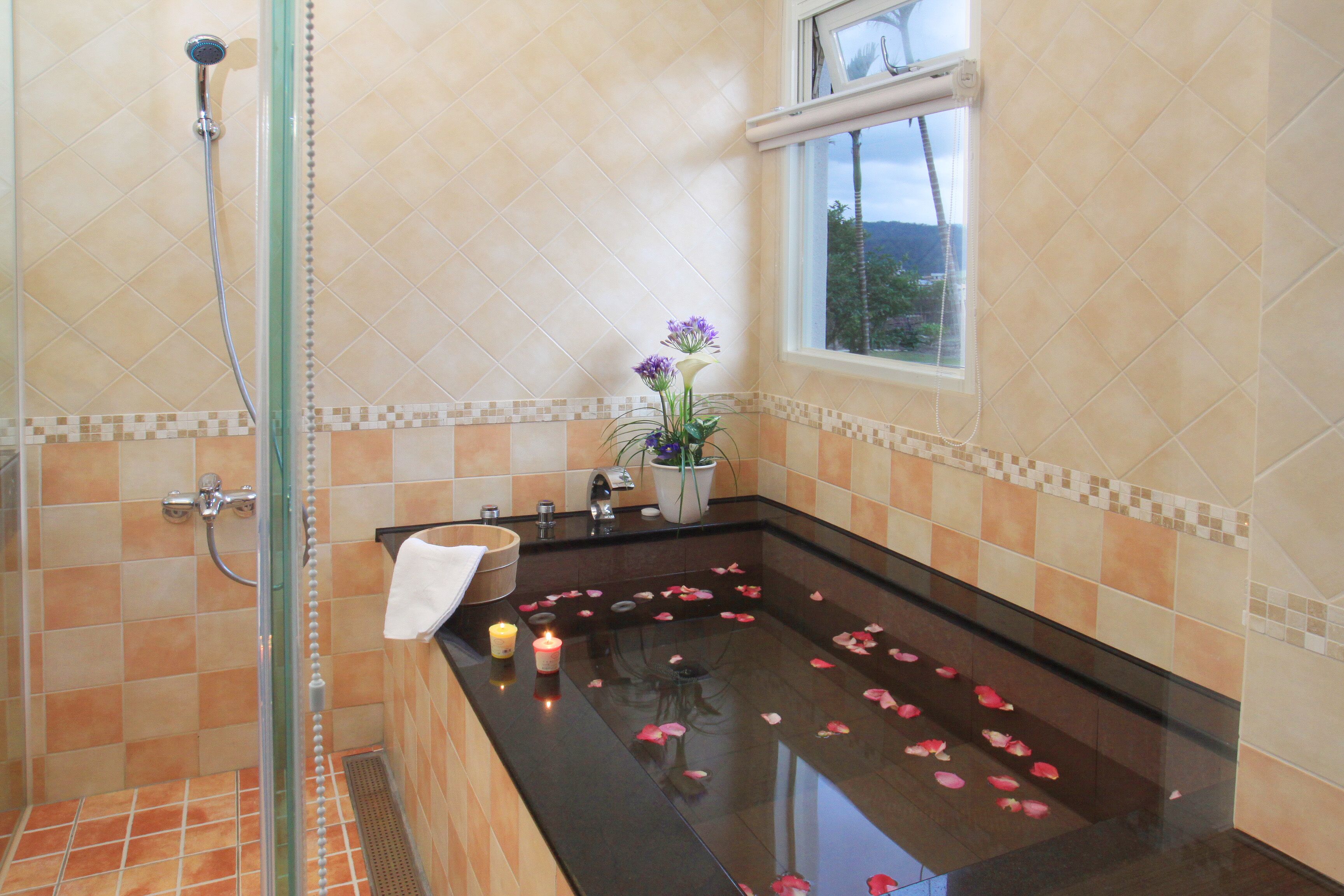 A bathroom with a glass-enclosed shower, a bathtub with rose petals, and a window with a view of greenery.