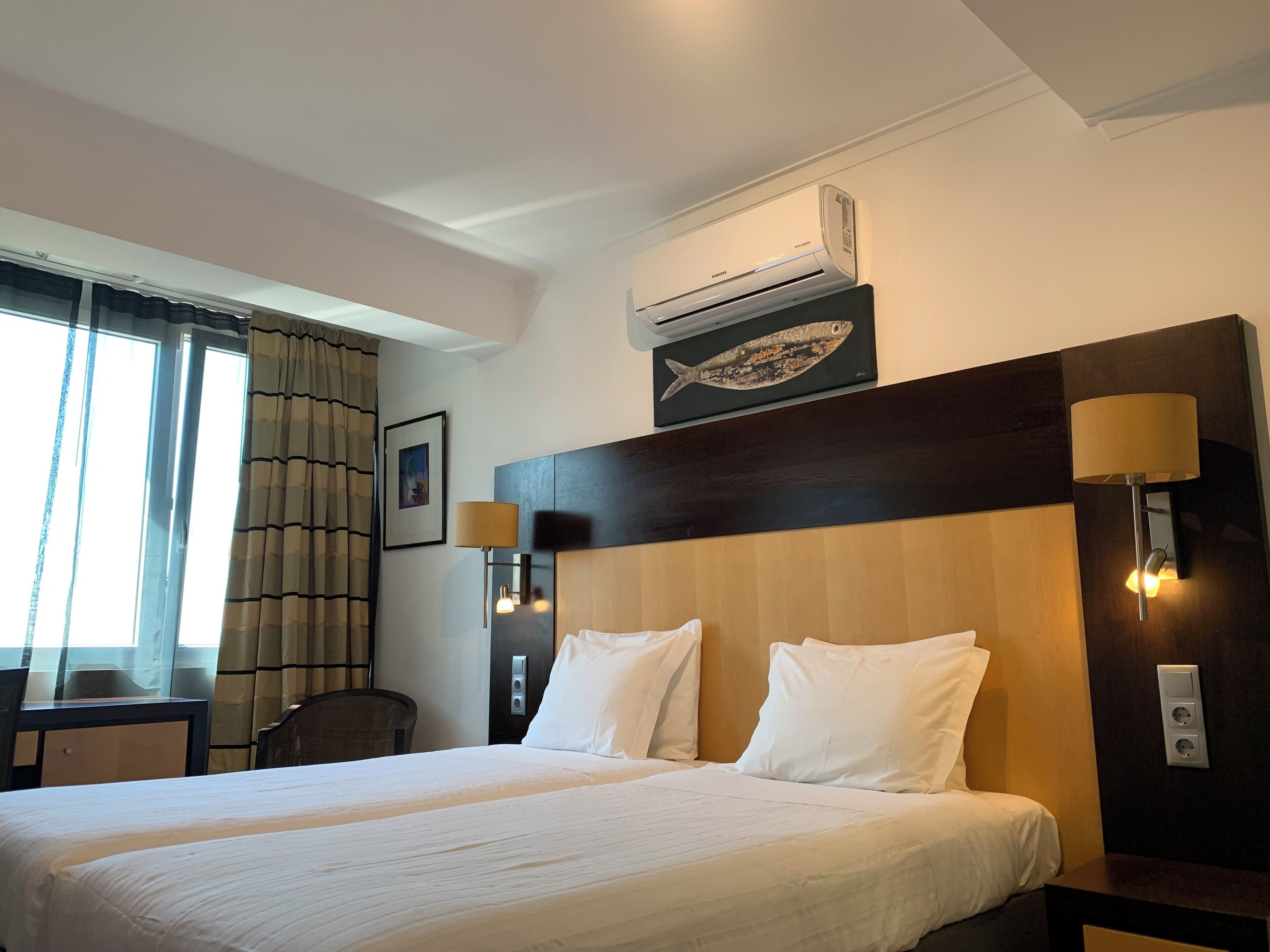 A hotel room with a large bed, a headboard with a fish painting, a window with curtains, a desk, and a wall-mounted air conditioner.