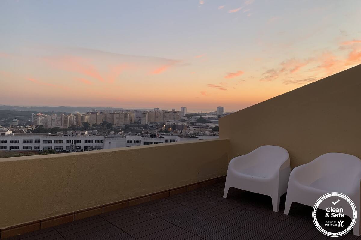 Ein Balkon mit zwei weißen Stühlen, Blick auf die Stadt und einem Sonnenuntergangshimmel.