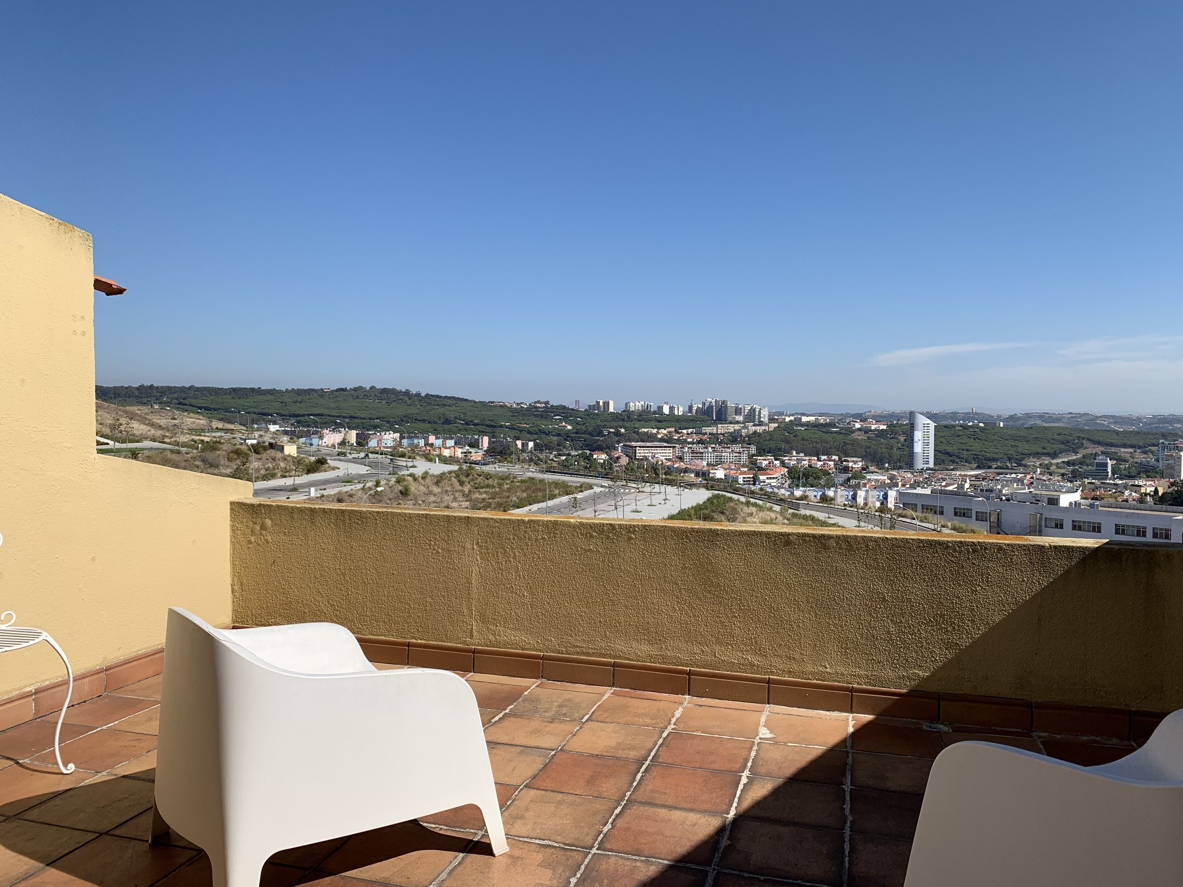 A terrace with two white chairs, a view of a cityscape with buildings and a hill, and a clear blue sky.