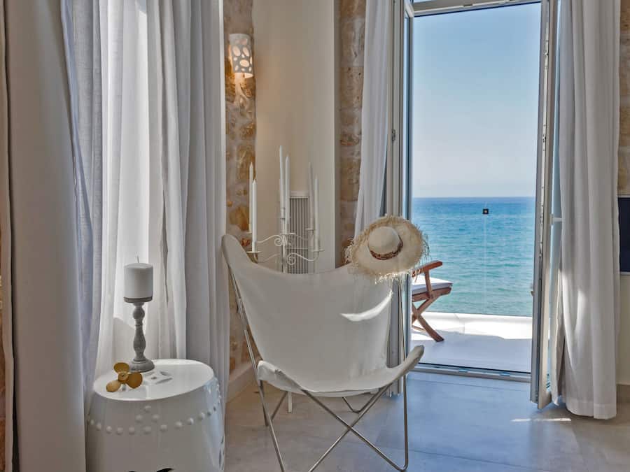 A room with a white chair, a small table with a candle, and a view of the sea through a glass door.