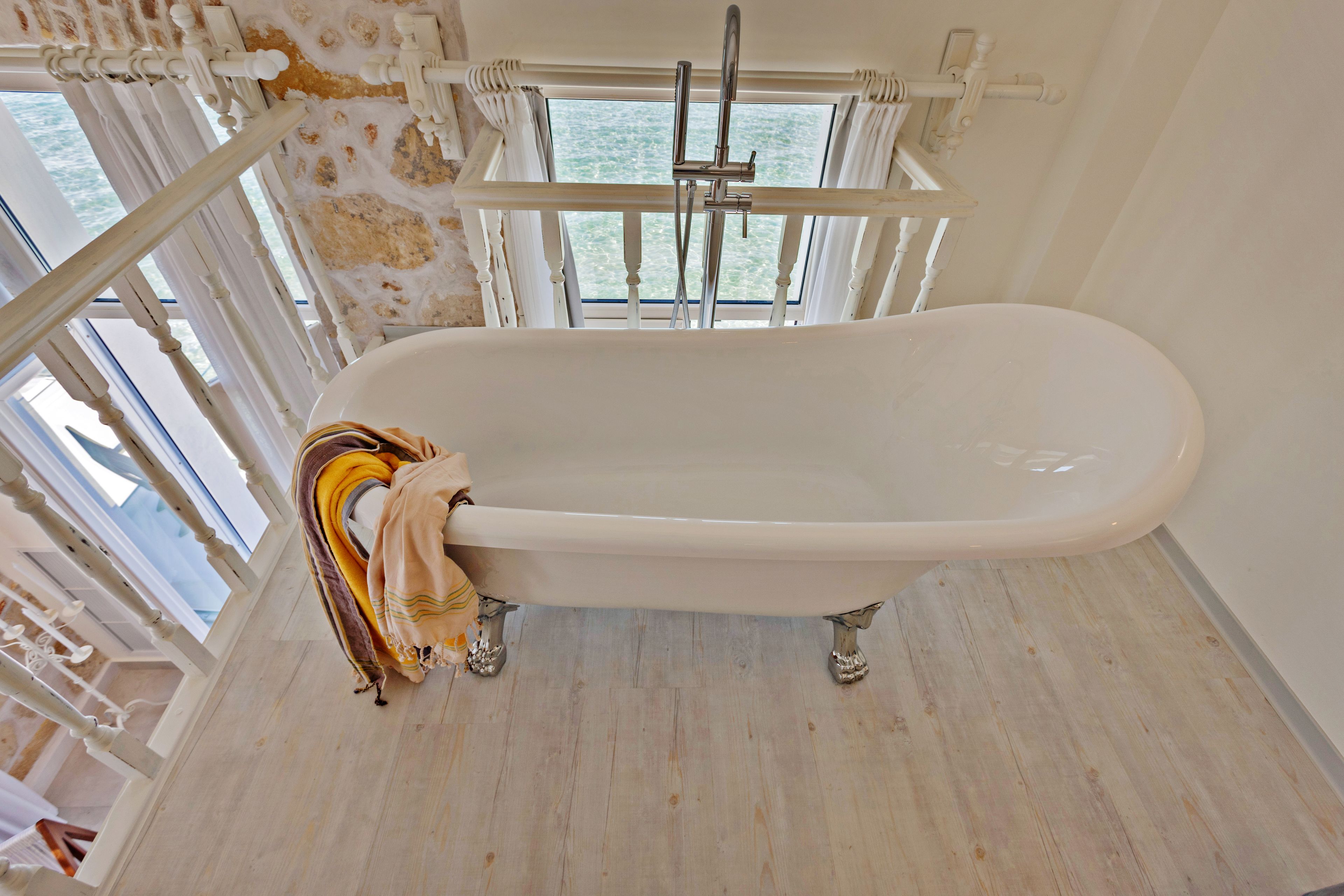A white bathtub with a showerhead in a room with a wooden floor and a window overlooking a body of water.