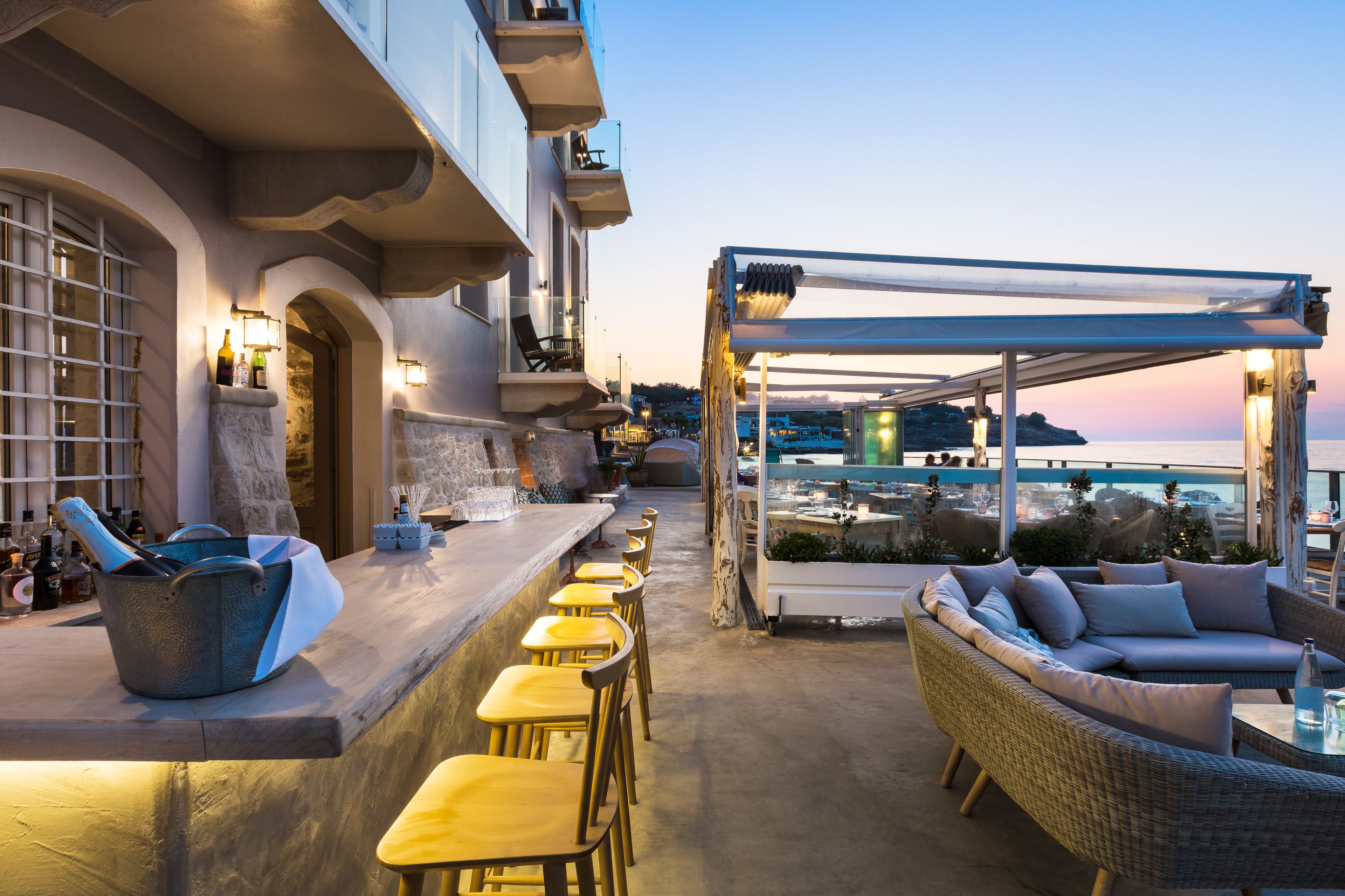A bar area with a long counter, yellow chairs, and a wicker sofa under a pergola.
