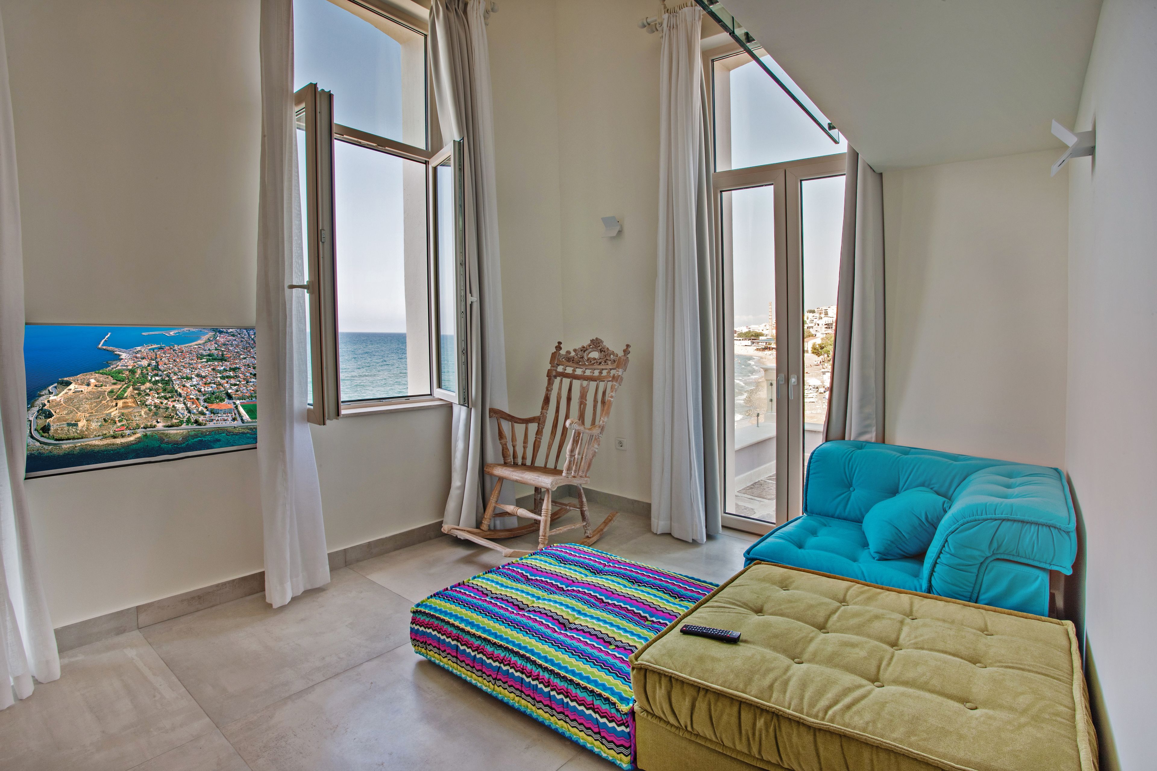 A room with a view of the sea, a rocking chair, a colorful striped ottoman, and a teal sofa.