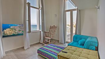 A room with a view of the sea, a rocking chair, a colorful striped ottoman, and a teal sofa.