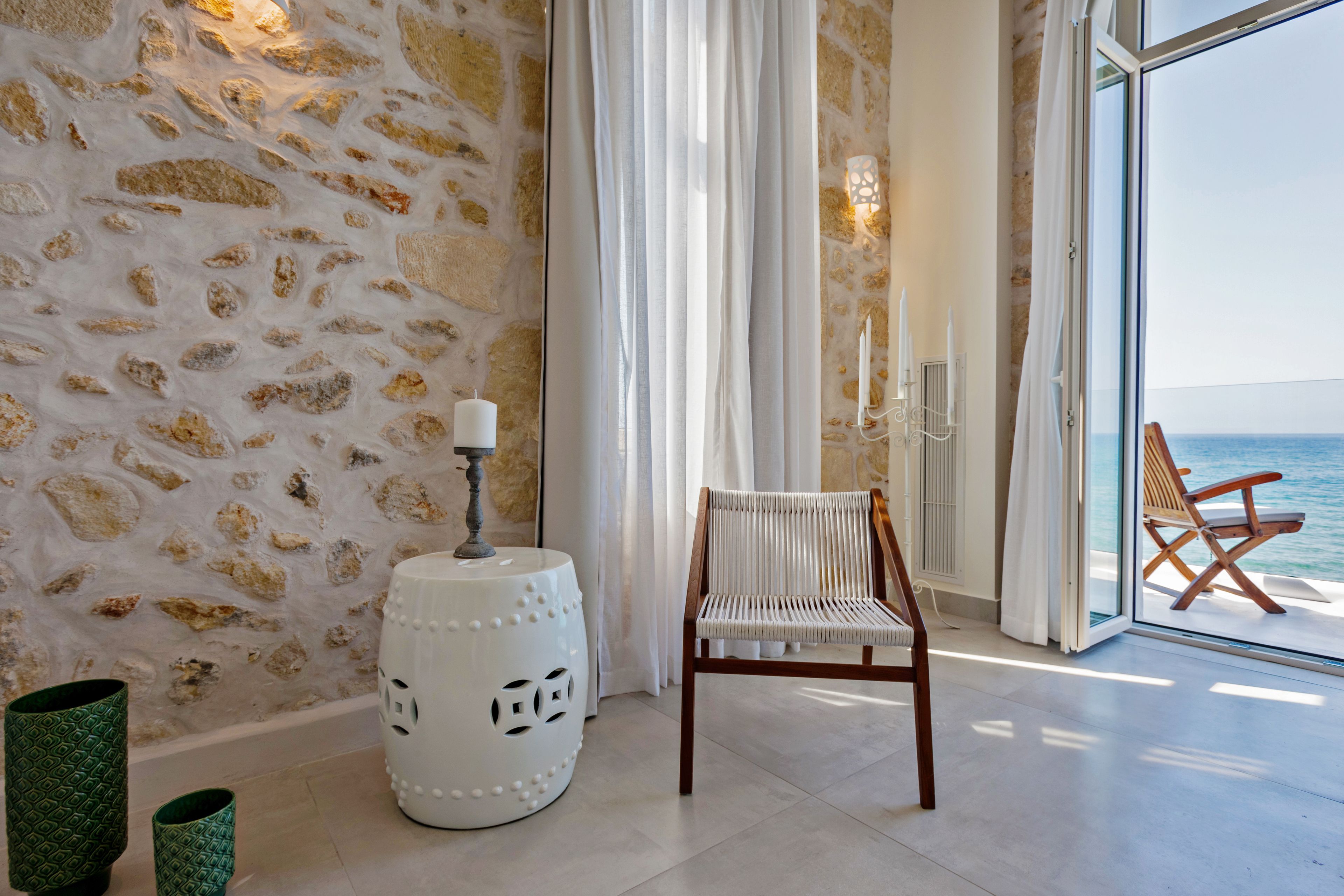 Une pièce avec un mur en pierre, une chaise blanche, une petite table blanche sur laquelle se trouve une bougie, et une vue sur la mer par une porte vitrée.