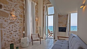 Amplia sala de estar con pared de piedra, un ventanal con vista al mar, un sofá gris, una silla de madera y una mesita.