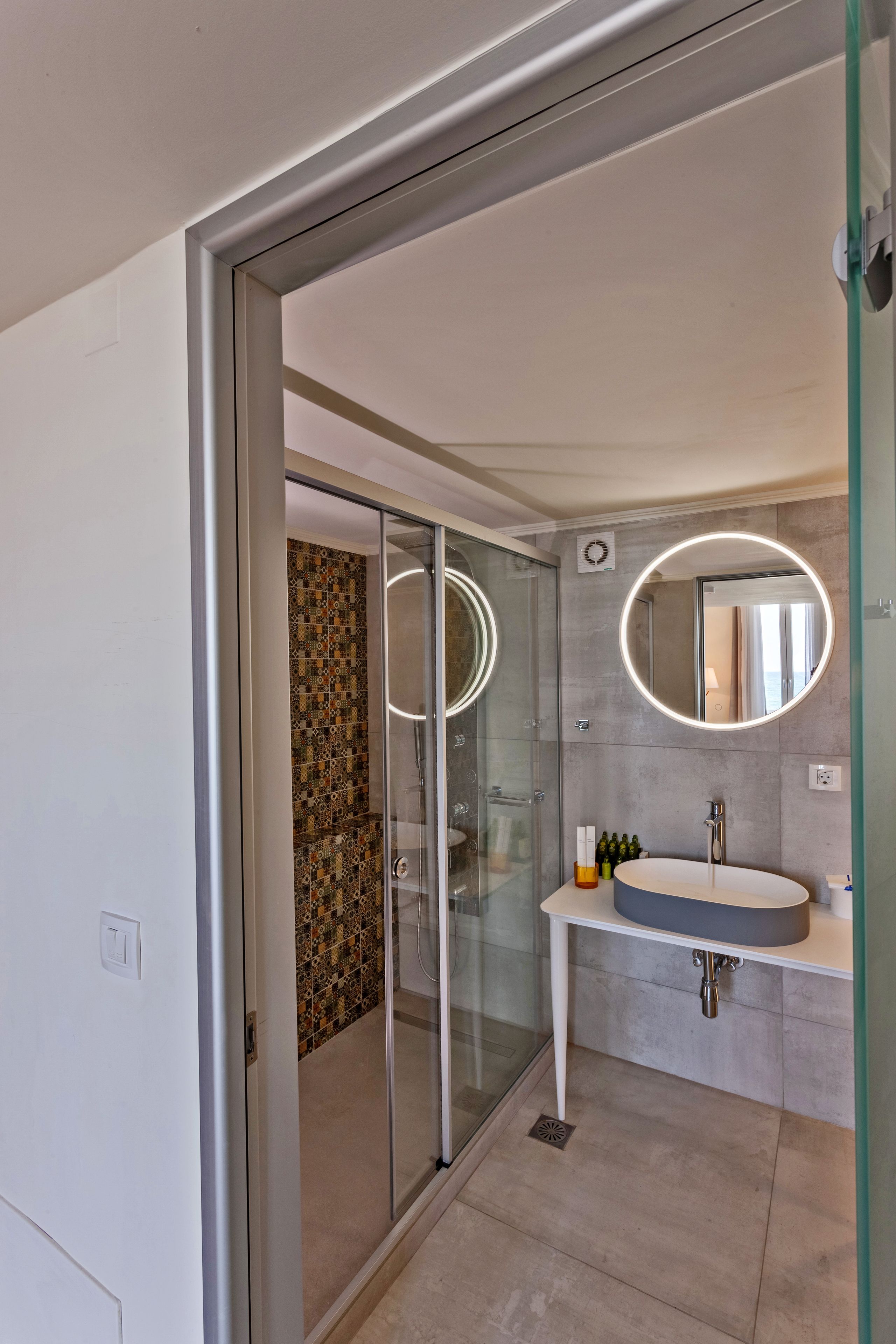 A modern bathroom with a glass shower enclosure, a round mirror, and a white sink.