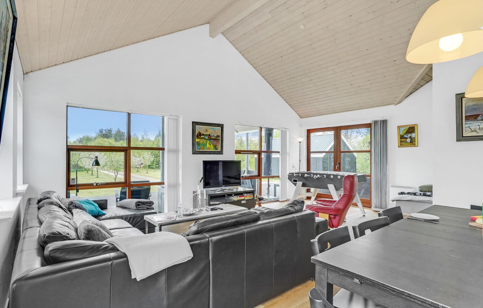 A spacious living area with a large sofa, a dining table, and a foosball table. There are large windows providing natural light and views of the outdoors.