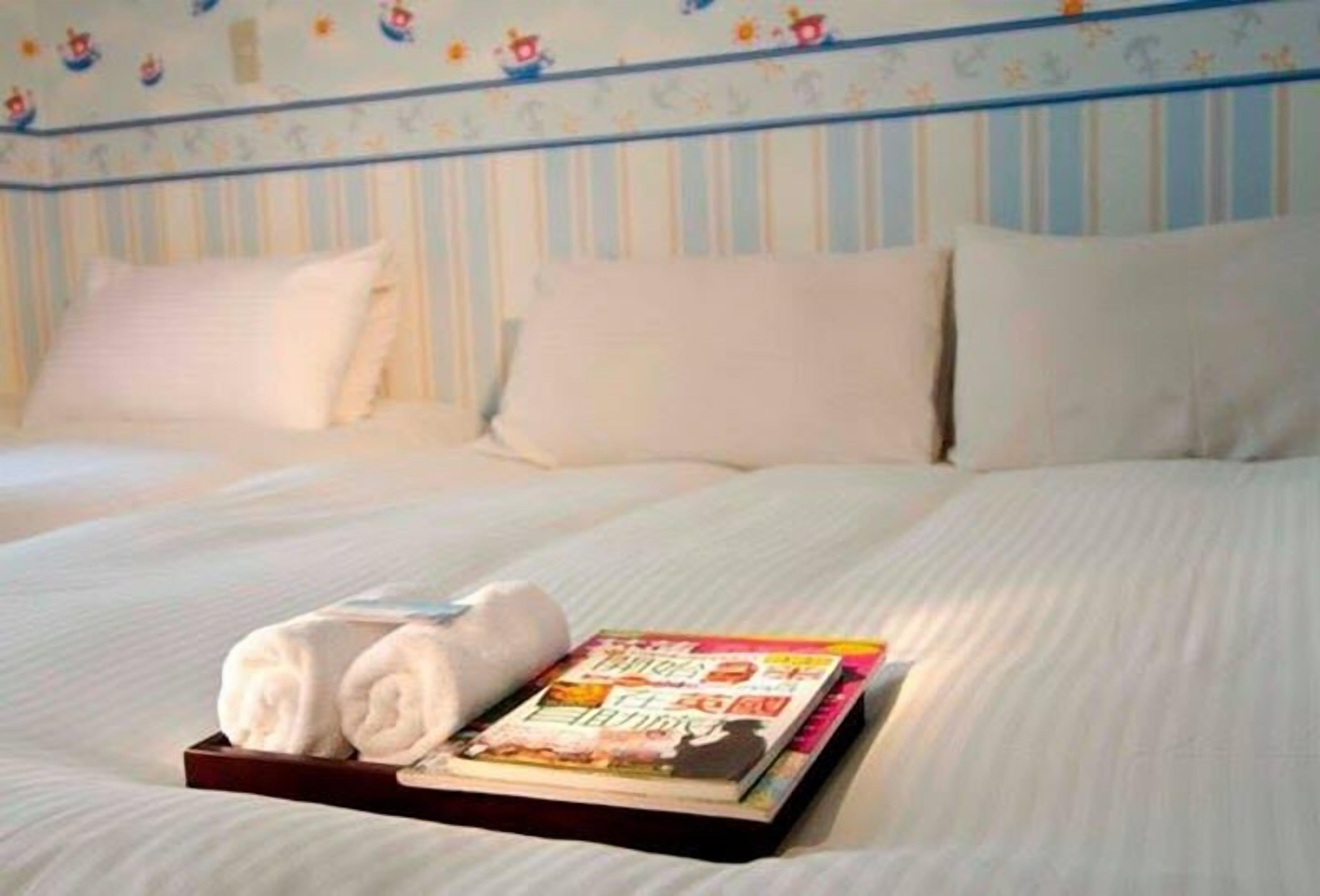 A neatly made bed with white linens and pillows, a rolled towel, and a book on a tray.