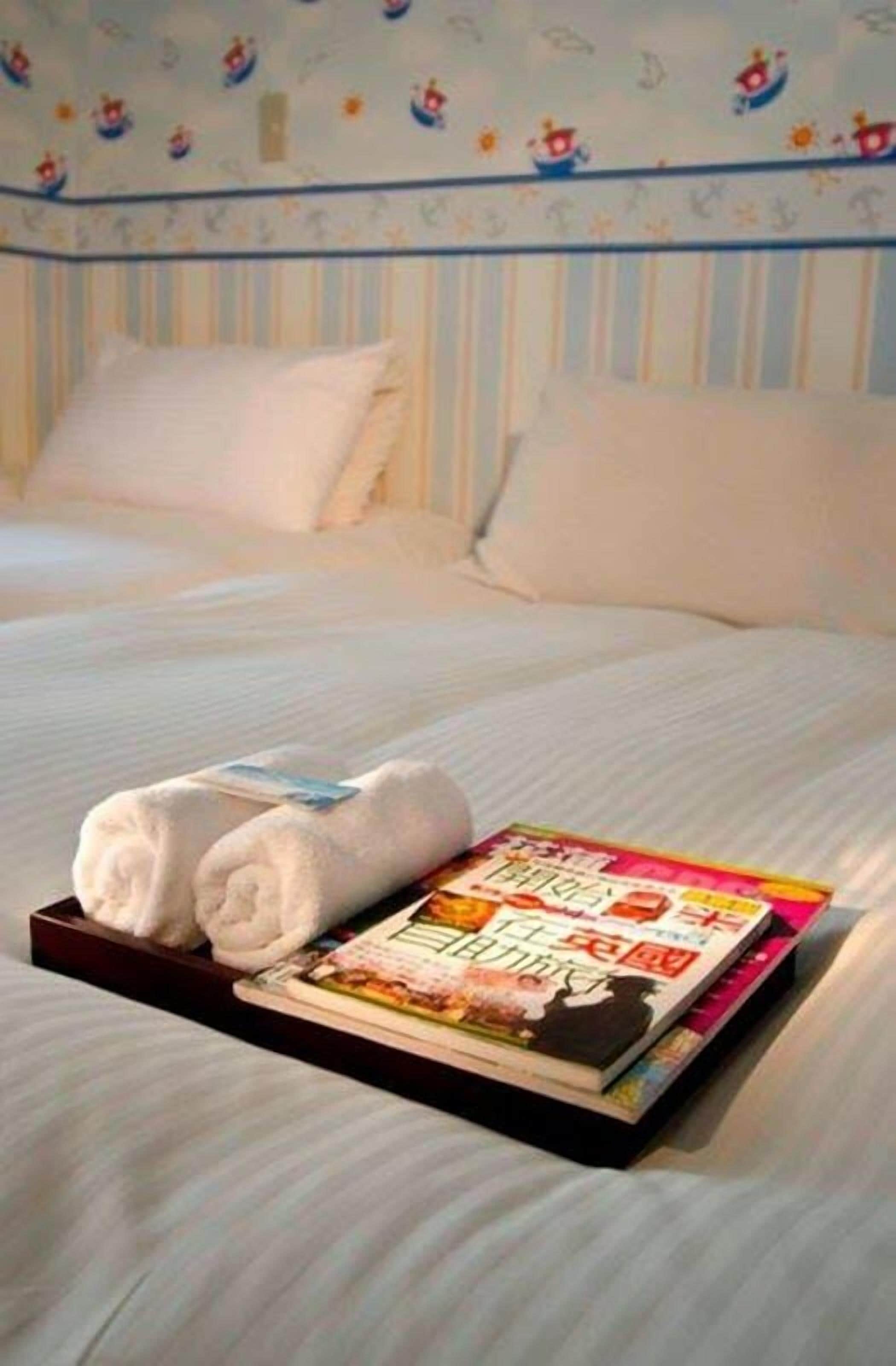 A neatly made bed with white linens, a folded towel, and a stack of books on a tray.