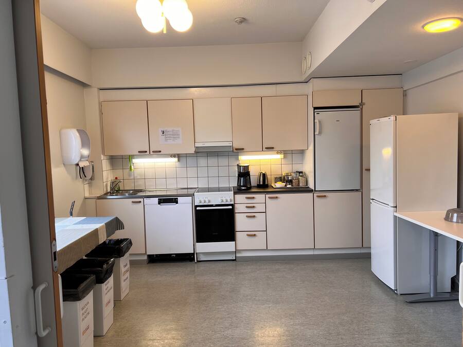 Cocina compacta con gabinetes blancos, estufa, refrigerador y cafetera. Hay botes de basura y un dispensador de papel en la pared.