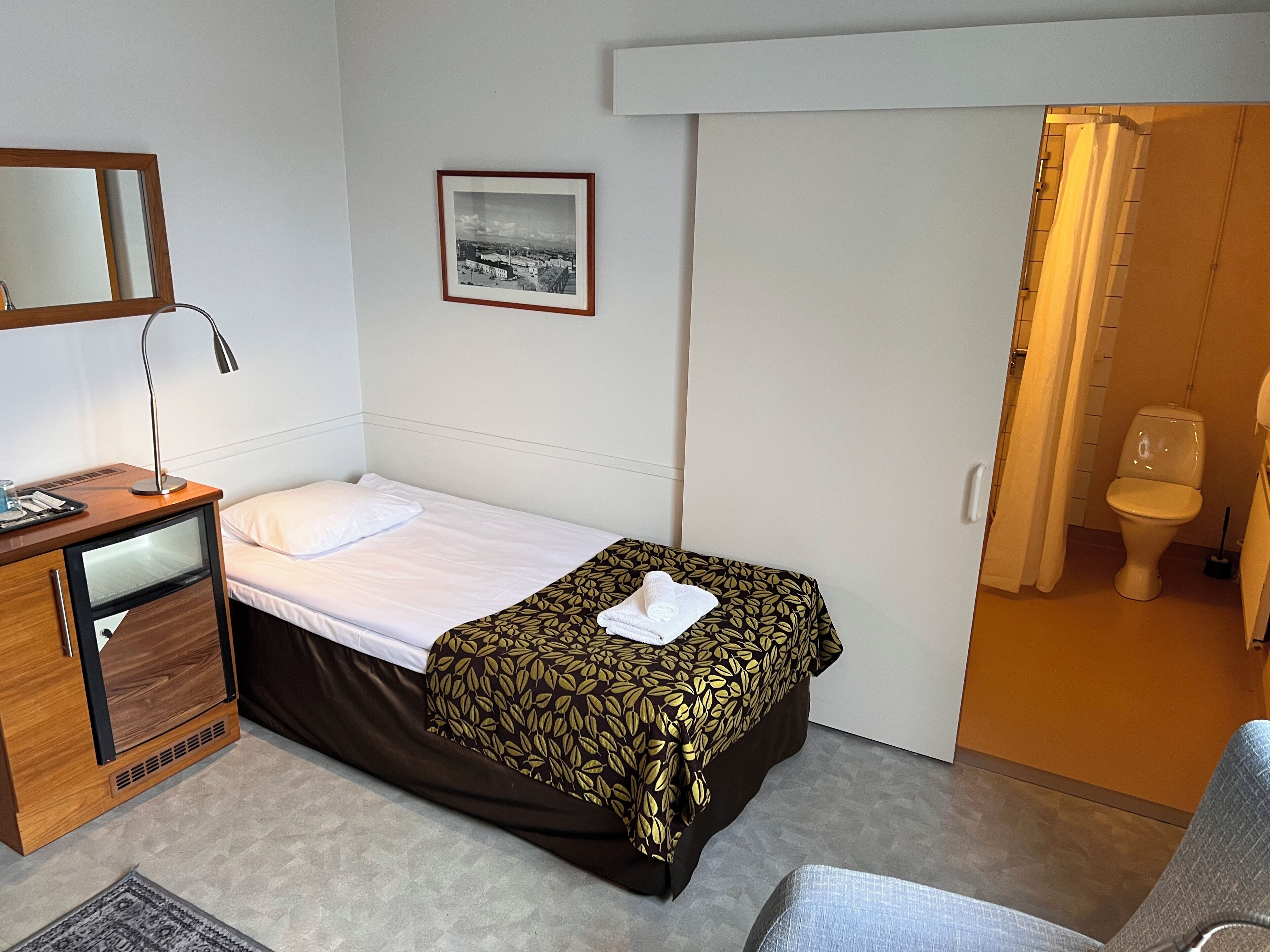 Habitación de hotel con cama, escritorio con lámpara, una alfombra pequeña y baño visible a través de una puerta abierta.